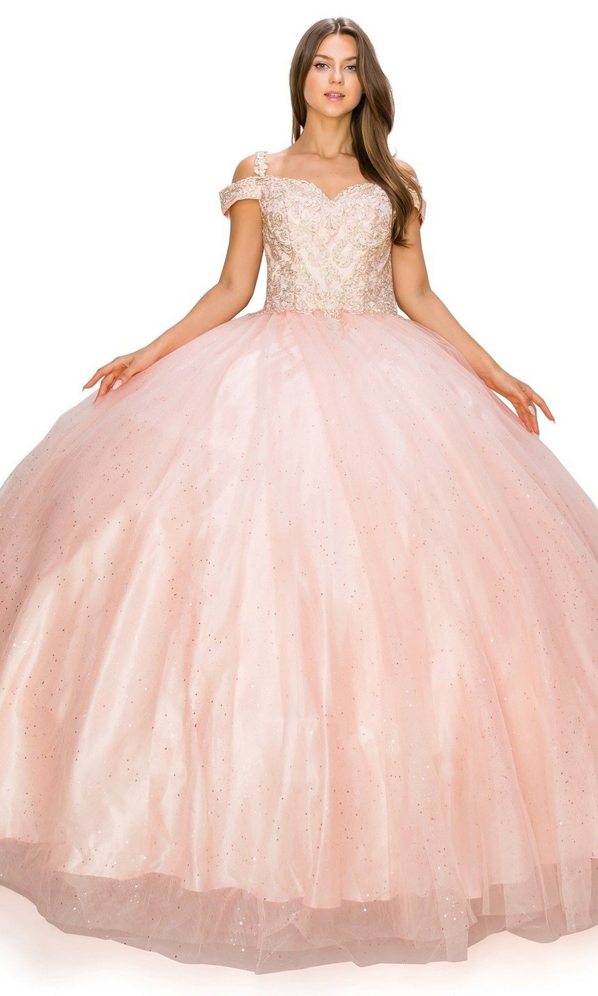 A woman stands against a plain background wearing the Quinceanera Ball Gown 8028J—an off-the-shoulder, light pink gown with a full, glitter tulle ball skirt.
